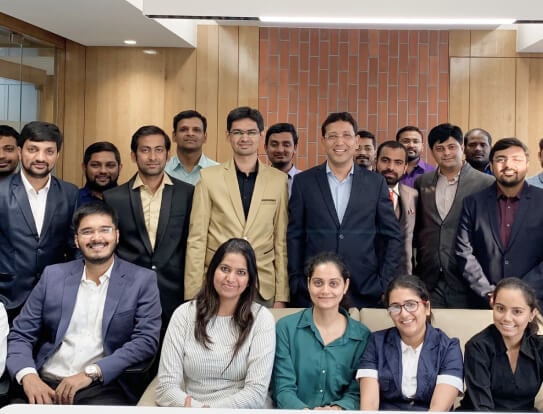 A group of Aakash colleagues seated and standing in front of a brick wall in the office.