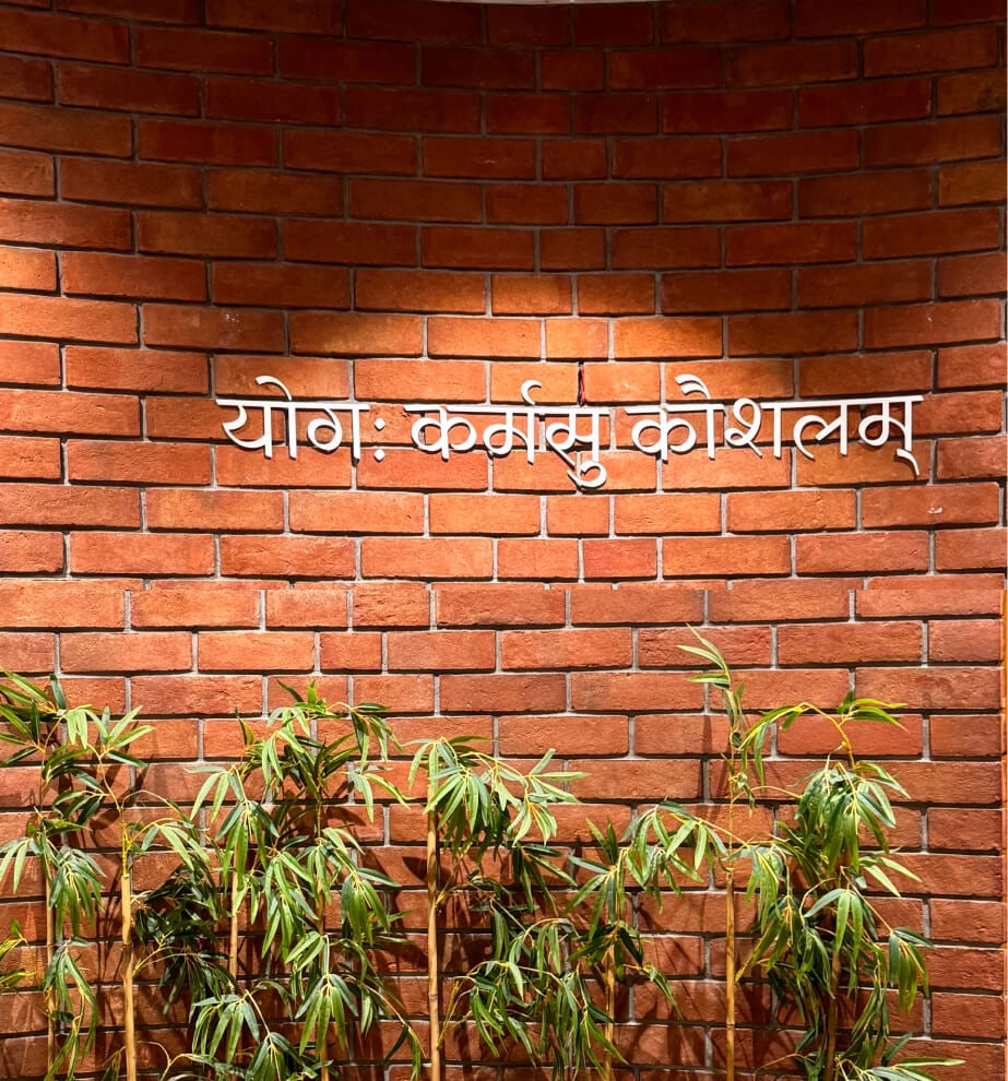 A red brick wall in Aakash's Ahmedabad office, with the Sanskrit line योगः कर्मसु कौशलम् mounted in white letters above a row of bamboo.